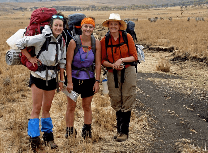 Brenna poses with two classmates. They're all wearing their hiking backpacks. In the background we see a herd of zebra.