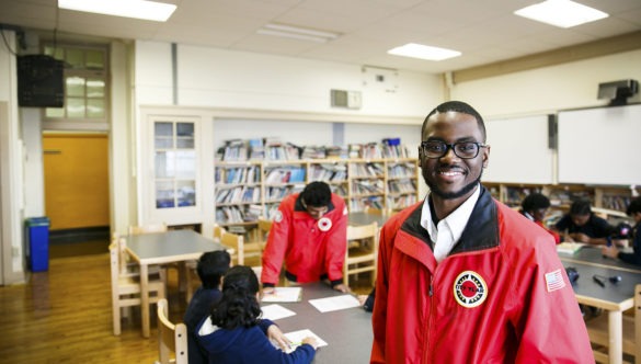 A City Year AmeriCorps member stand in a library, looking proudly at the camera. Behind him are students working on homework with another AmeriCorps member