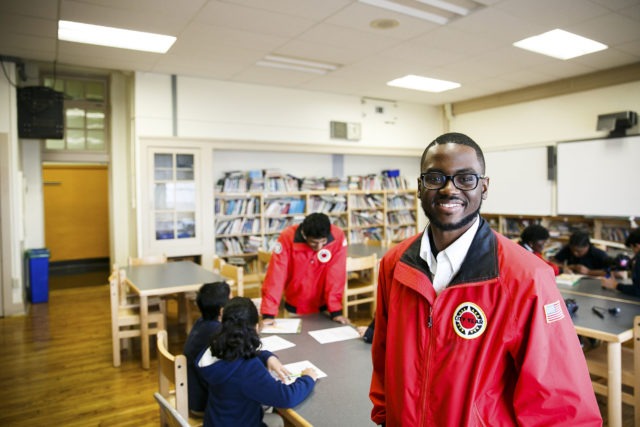 A City Year AmeriCorps member stand in a library, looking proudly at the camera. Behind him are students working on homework with another AmeriCorps member