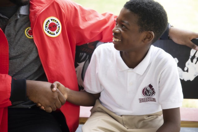 a young student smiles and looks up at an AmeriCorps member while shaking their hand