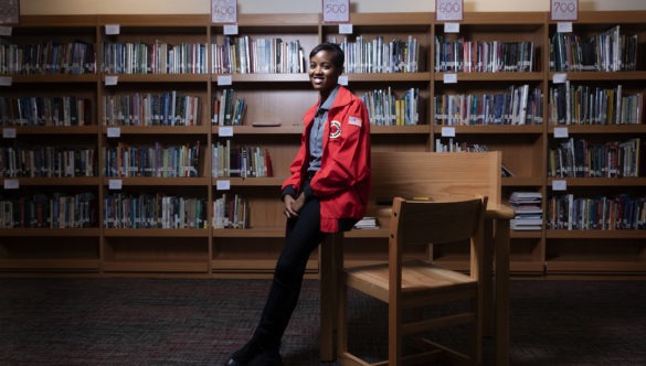 AmeriCorps member leaning on table smiling in the school library with shelves of books in background