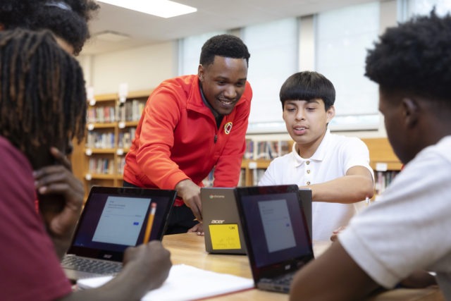 three students are working at computers in the library, while an AmeriCorps member works with one of the students