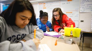 an AmeriCorps member is leaning over to point out something on a student's paper while they work on an assignment in class