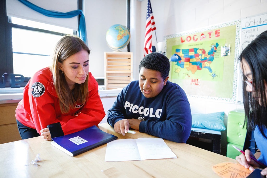 An AmeriCorps member leans on a desk, looking at a worksheet with a student