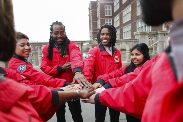 A team of nine AmeriCorps members stand with their hands in a the center of a circle, ready to do a spirit break