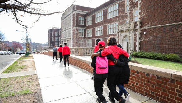 Two students with backpacks walking on a sidewalk near a school with a City Year Corps Member