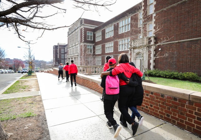 Two students with backpacks walking on a sidewalk near a school with a City Year Corps Member