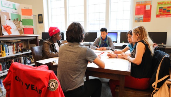 Four students and a City Year corps member learning at a large table in a classroom with computers and bookshelves