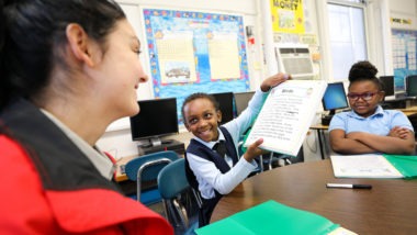 a young student holding up a page in a book to show a city year corps member while another student looks on.