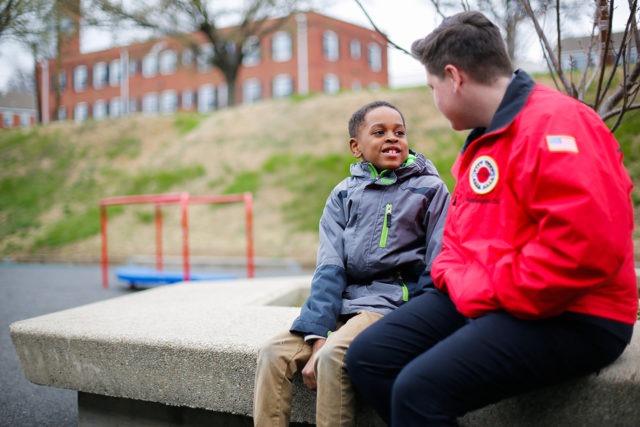 A young student in a jacket sitting with a city year corps member in a school playground in the winter