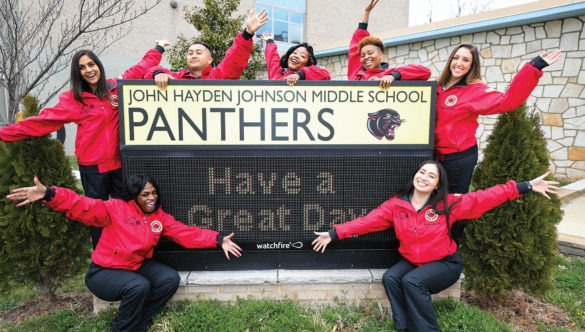 AmeriCorps members wave their hands in front of the entrance sign to a middle school.