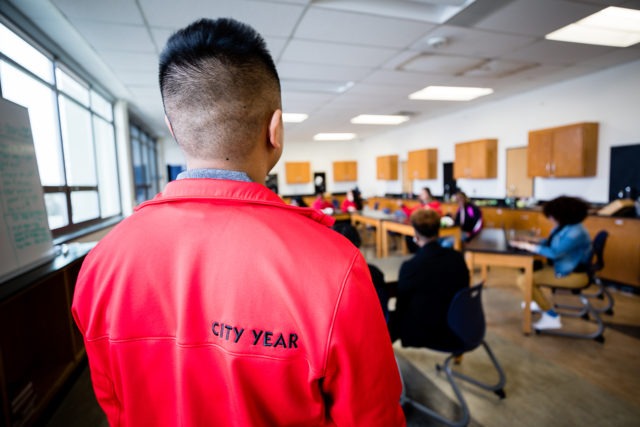 the back of a city year americorp member with the label City Year on his jacket as he looks over a teacher and students in a classroom