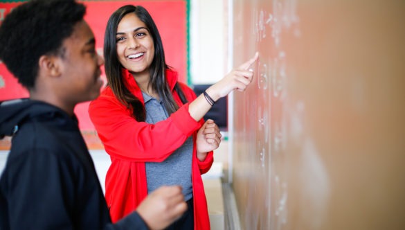 A city year americorps member pointing out an answer to a math problem on a blackboard as a student looks at the work