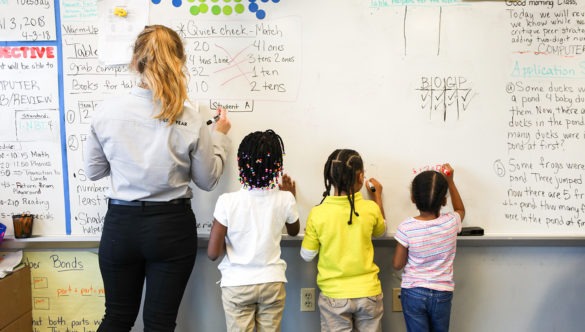 The back City year americorps member standing next to three young students all facing the white board they are writing on