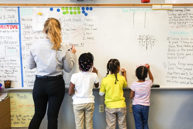The back City year americorps member standing next to three young students all facing the white board they are writing on