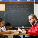 City year americorps member reading along with two students as they sit at desks in a classroom