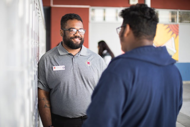 city year americorps member talking with a high school student as they stand in front of lockers in a school hallway