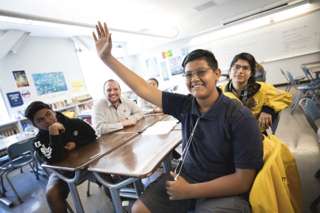 A student confidently raises his hand in class while his proud AmeriCorps members sit behind him
