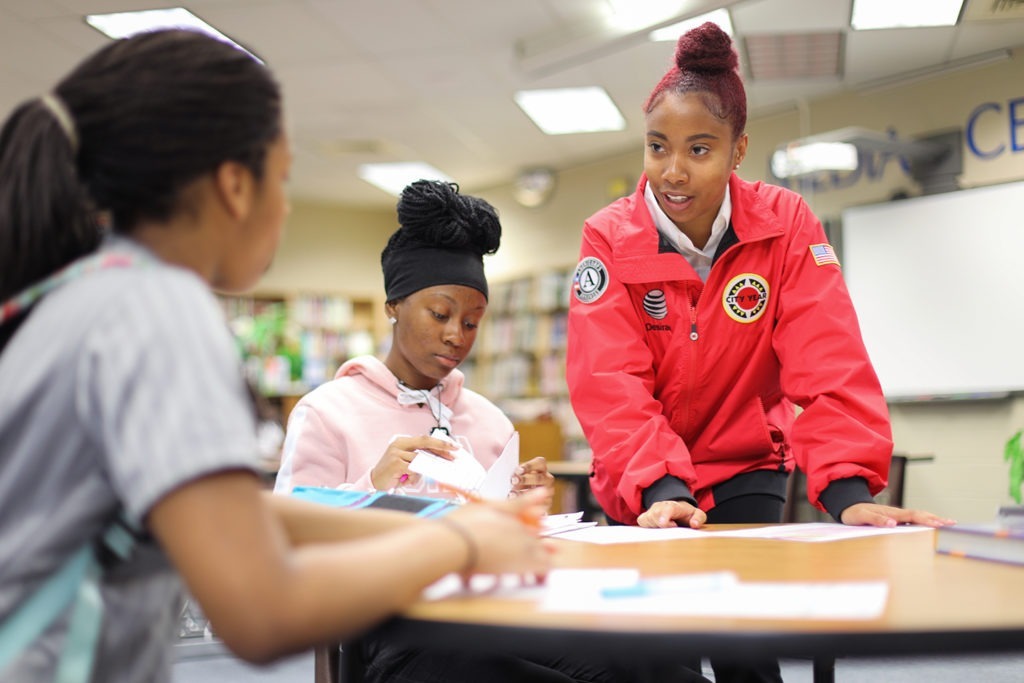 city year americorps member stands and helps two students who are sitting at a table working on schoolwork