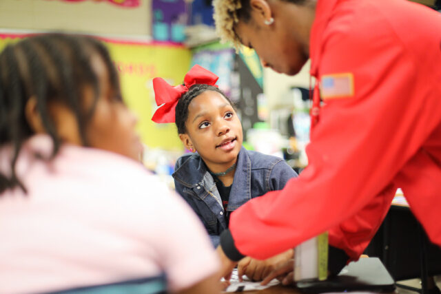 a student looks up at an AmeriCorps member as they help them at their desk