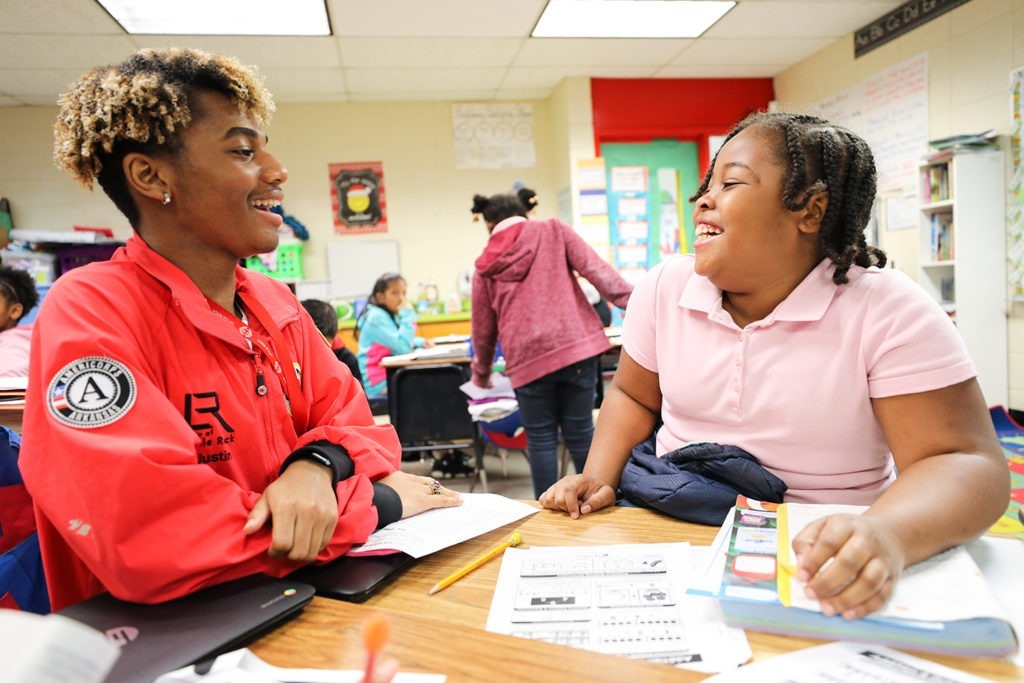 AmeriCorps member and student are smiling and laughing together as they do work at a table in the classroom