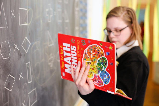 city year americorps member reading a book titled math genius while standing in front of a chalkboard