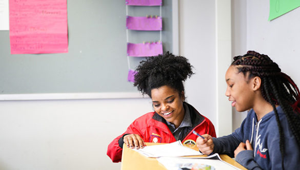 City Year AmeriCorps member kneeling next to a student at a desk as they review work