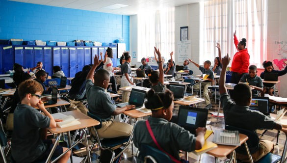 Students sitting at desks in a classroom with computers in front of them some raising hands as a City Year AmeriCorps member stands with her hand raised
