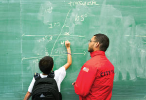 City year americorps member and a student with a back pack at a classroom chalkboard as the student solves math problems