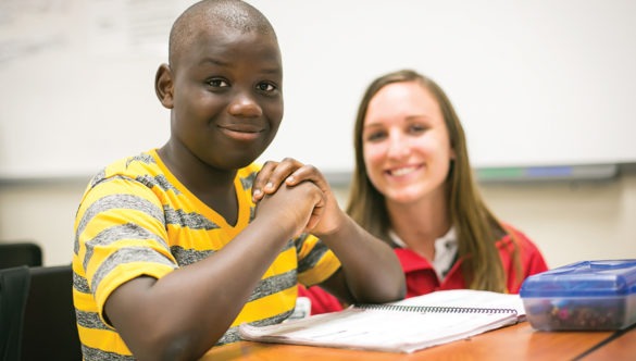 A student smiling for the camera in a classroom while a City year americorps member looks at the camera in the background