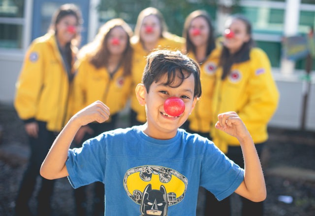 A young student flexes their muscles while wearing a Red Nose. Behind him, blurred in the background, are five AmeriCorps members also supporting Red Nose Day