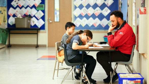 Two students sitting at a small desk doing their work as a City year americorps member sits facing them and watching what they are doing