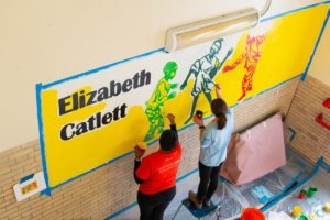 Two volunteers paint a mural of three children playing soccer together in a school stairwell