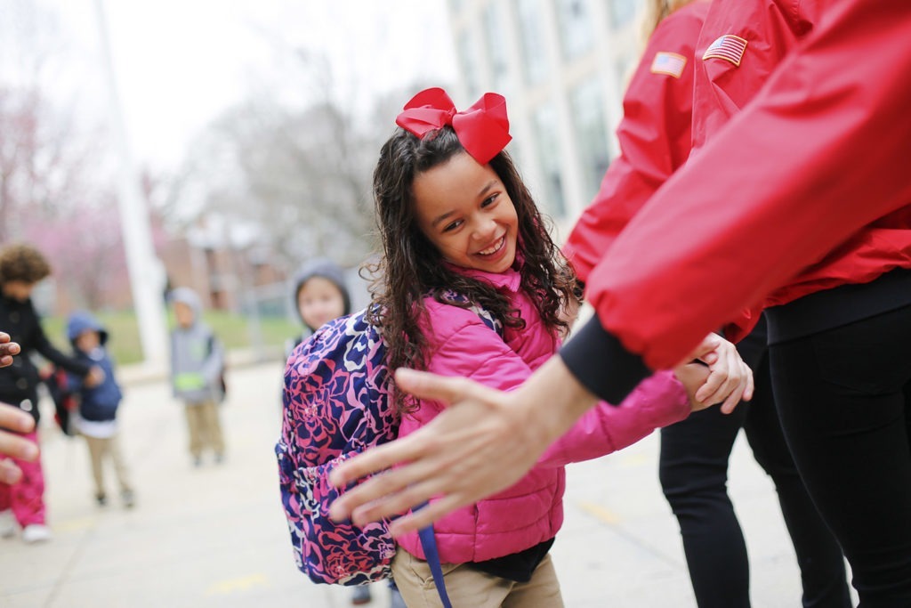 An elementary school student wearing a bow and a backpack high-fives several AmeriCorps members as she arrives at school on time