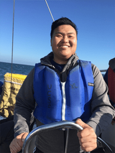JOhn Yu smiles while standing at the wheel of a sailboat.