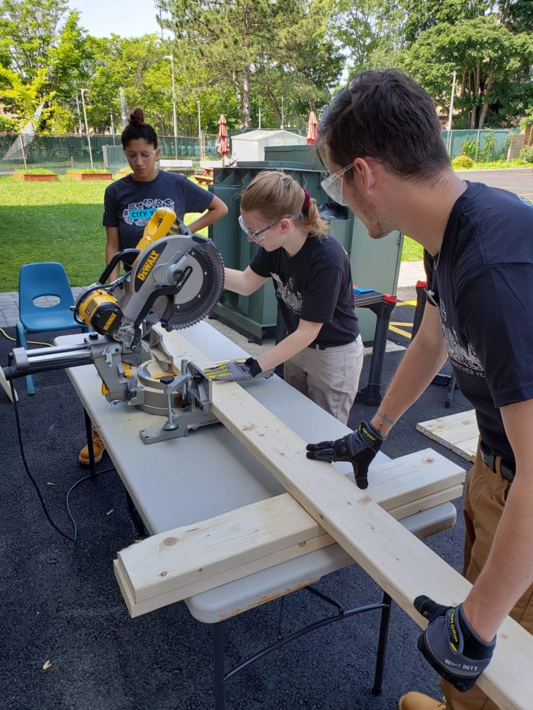 Three Care Force members work together to cut wood using a chop saw. One member operates the saw while another supports the longer end of wood. The third Care Force member observes the process.