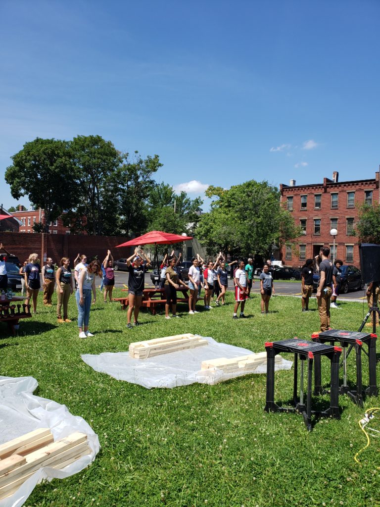 WilmerHale volunteers participate in physical training (PT) with Team Care Force. Bench kits are laid out of tarp in front of them while they perform Peel Bananas.