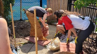 WilmerHale volunteers and Care Force member work on a paver stone path between the sidewalk and the gate to the playground. There is a wheelbarrow in the bottom right corner and a tamper standing next to it.