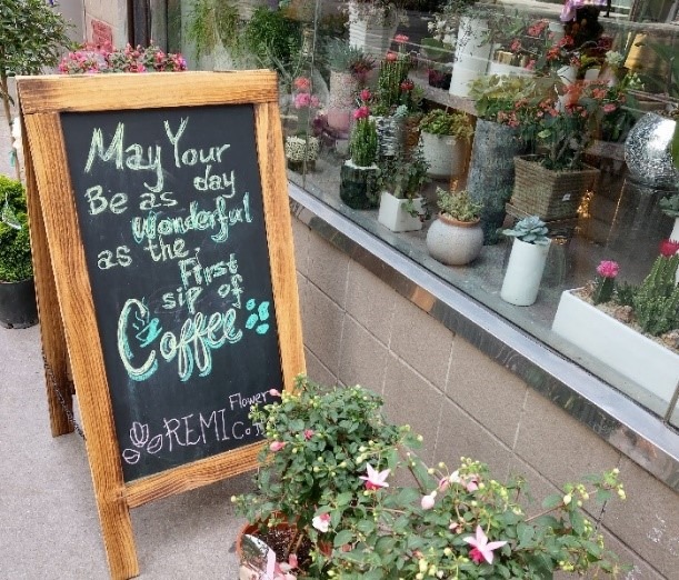 A chalkboard sign with the words "May your day be as wonderful as the first sip of coffee" sits next to a ledge covered with various types of plants.