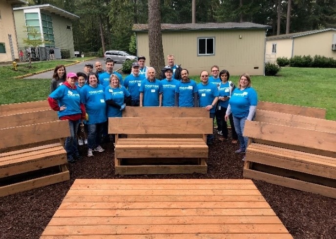 A group of AT&T volunteers wearing blue t-shirts stand around benches with backs and a stage.