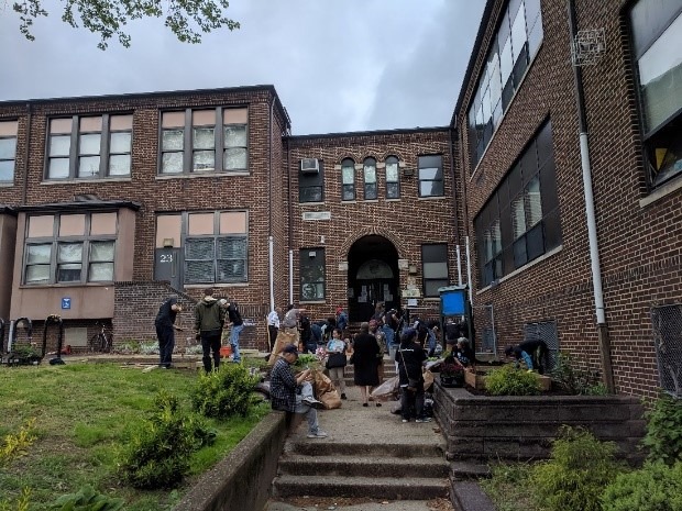 AT&T volunteers in blue shirts construct garden beds and benches in front of the Rivera Community Middle School entrance on a cloudy day.
