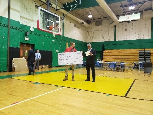 Senior AmeriCorps Member Clark Shimeall stands holding a large check in his iconic red jacket next to a Taco Bell representative after receiving the Taco Bell Live Más Scholarship.