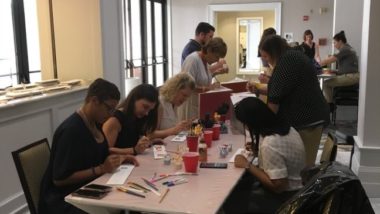 Volunteers are being led in decorating colorful mural tiles with words from the books that HMH donated to the school. Also volunteers in the back are decorating school emblems with the school colors on the roofs of the free little libraries.