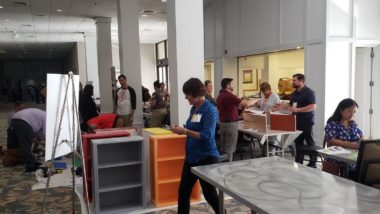 Volunteers are making origami bookmarks to the front right of the photo. To the middle left volunteers are working on constructing free little libraries. To the middle right volunteers are working on decorating the school emblems and in the back volunteers are working on decorating the mural tiles for the sides of the free little libraries.