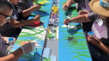 Volunteers sit at a picnic table painting bugs and grass onto bench backs.