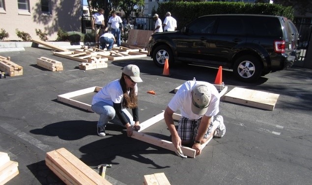 One man and one woman both wearing hats and white shirts hold planks of wood in place.