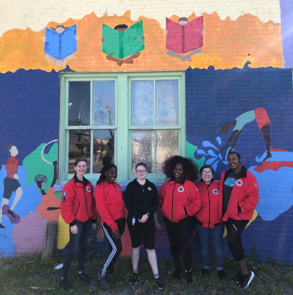 AmeriCorps members stand in front of a colorful mural with their red jackets on.