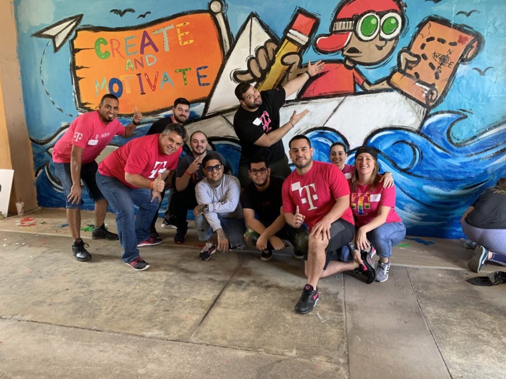 T-Mobile volunteers pose in front of mural of a young boy in a boat with a pancil and map in hand. His flag has the phrase Create and Motivate written on it in different colors.