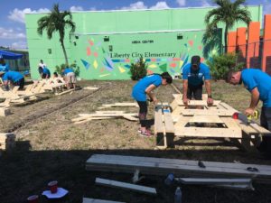 Volunteers in blue shirts build adult picnic tables on the lawn of Liberty City Elementary School.