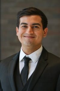 Headshot of Care Force alumni Alejandro wearing a black suit and tie standing in front of a gray background.